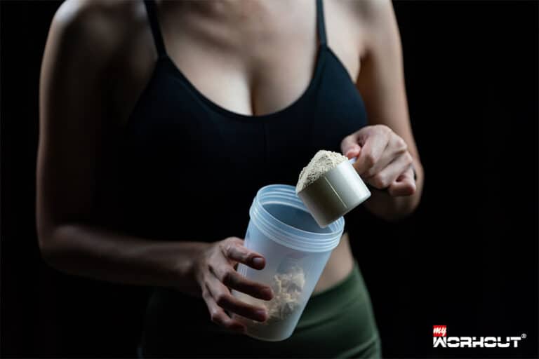 Vegane Proteinshakes - Wie gesund sind sie wirklich? Frau im Sportbh hält einen Becher mit Proteinpulver vor einem schwarzen Hintergrund